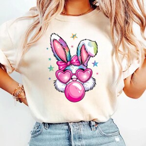 May include: A white t-shirt with a colorful cartoon bunny wearing pink sunglasses and blowing a bubble. The bunny has a pink bow on its head and is surrounded by stars.