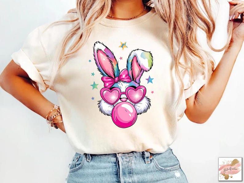 May include: A white t-shirt with a colorful cartoon bunny wearing pink sunglasses and blowing a bubble. The bunny has a pink bow on its head and is surrounded by stars.