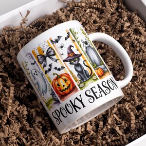 May include: White ceramic mug with a Halloween-themed design. The mug features illustrations of a ghost, bats, a jack-o'-lantern, a black cat in a witch hat, and a tombstone. The words "Spooky Season" are printed on the mug.