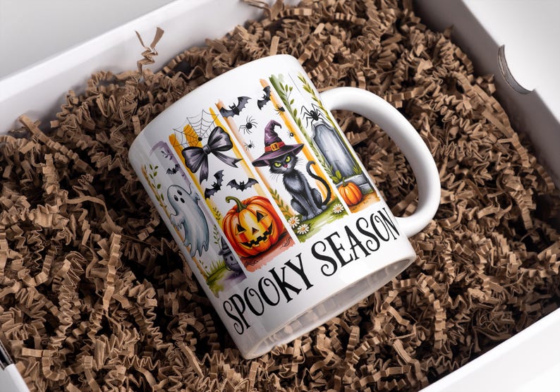 May include: White ceramic mug with a Halloween-themed design. The mug features illustrations of a ghost, bats, a jack-o'-lantern, a black cat in a witch hat, and a tombstone. The words "Spooky Season" are printed on the mug.