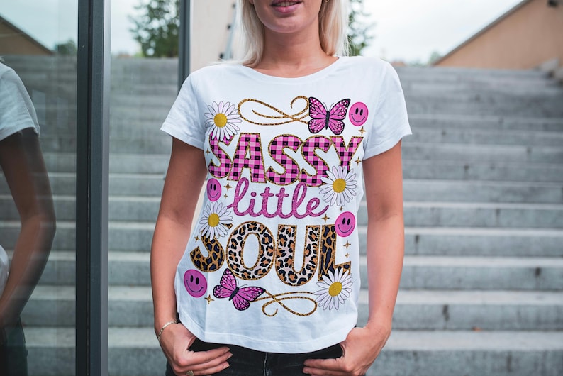 May include: White t-shirt featuring a pink and black checked design that reads "Sassy little soul". The design also incorporates butterflies, flowers, and smiley faces.