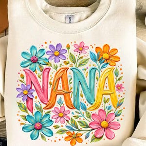 K&ouml;nnte beinhalten: Cremefarbenes Sweatshirt mit dem Wort "NANA" in bunten 3D-Buchstaben, umgeben von lebendigen floralen Designs in Rosa, Blau, Orange und Gelb. Die Blumen und Bl&auml;tter bilden ein fr&ouml;hliches, auff&auml;lliges Design.