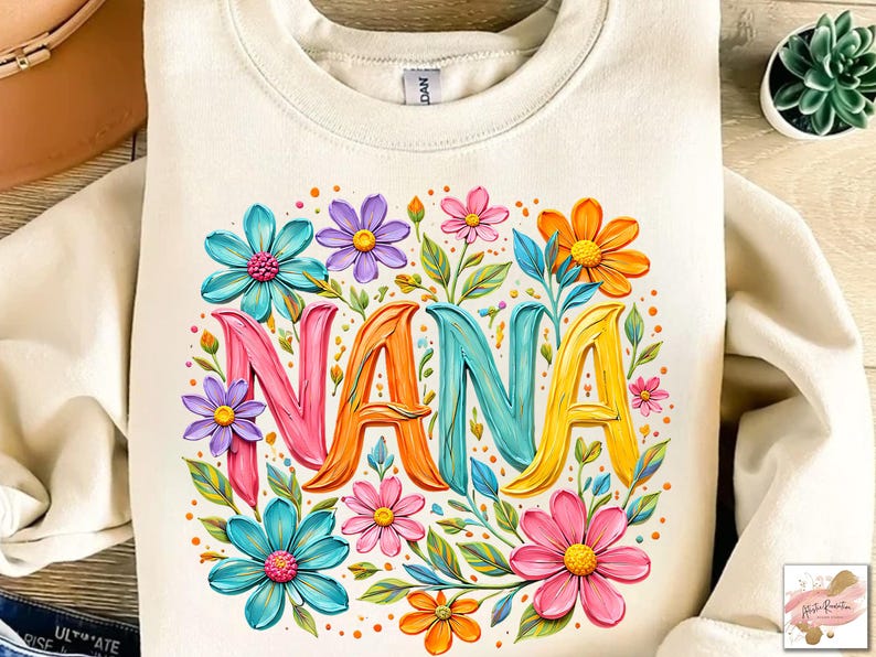 K&ouml;nnte beinhalten: Cremefarbenes Sweatshirt mit dem Wort "NANA" in bunten 3D-Buchstaben, umgeben von lebendigen floralen Designs in Rosa, Blau, Orange und Gelb. Die Blumen und Bl&auml;tter bilden ein fr&ouml;hliches, auff&auml;lliges Design.