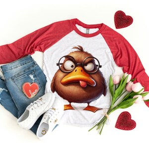 May include: A white and red baseball tee shirt with a brown cartoon duck wearing glasses and sticking out its tongue. The shirt is laying on a white surface with a pair of white sneakers, blue jeans, and a bouquet of pink tulips.