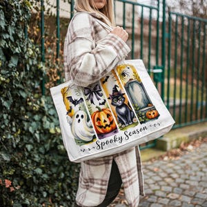 May include: White tote bag with a Halloween-themed design. The bag features a ghost, pumpkin, black cat, and tombstone. The words "Spooky Season" are printed on the bag.