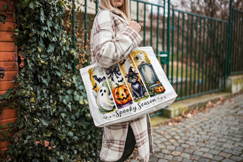 May include: White tote bag with a Halloween-themed design. The bag features a ghost, pumpkin, black cat, and tombstone. The words "Spooky Season" are printed on the bag.