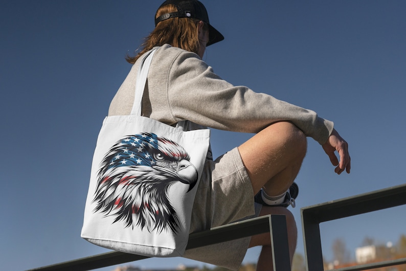 May include: White tote bag featuring a detailed illustration of an American bald eagle's head. The eagle's head is colored with the American flag design, including red, white, and blue. The bag has white straps and is carried over the shoulder.