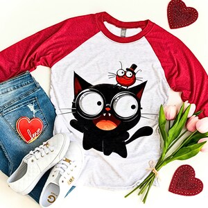 K&ouml;nnte beinhalten: Ein wei&szlig;es und rotes Baseball-T-Shirt mit einer schwarzen Katze, die eine Brille tr&auml;gt, und einem roten K&auml;fer mit einem Zylinder auf dem Kopf. Das Shirt liegt auf einer wei&szlig;en Oberfl&auml;che mit einem Paar wei&szlig;en Sneakers, einer blauen Jeans und einem Strau&szlig; rosa Tulpen.