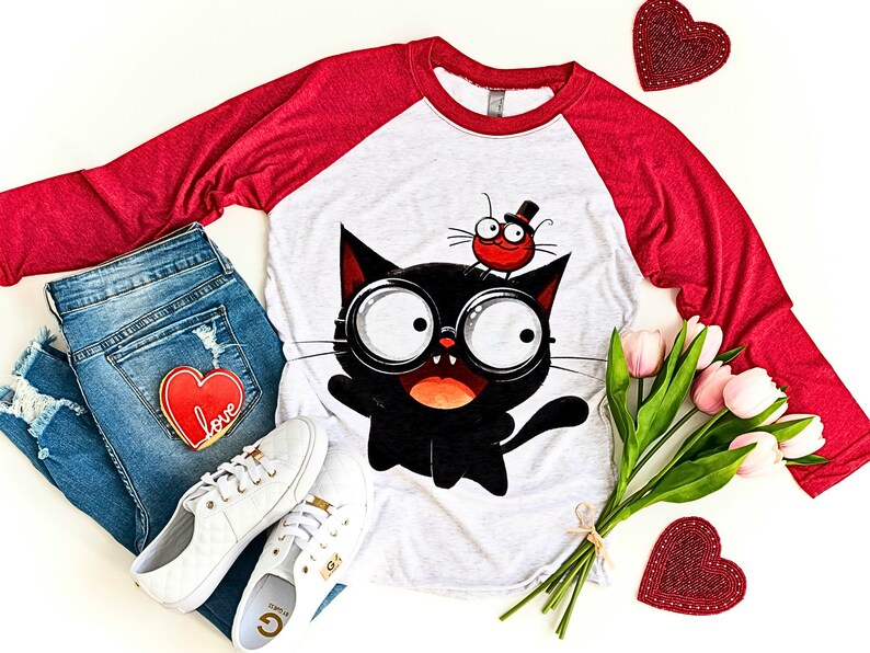 K&ouml;nnte beinhalten: Ein wei&szlig;es und rotes Baseball-T-Shirt mit einer schwarzen Katze, die eine Brille tr&auml;gt, und einem roten K&auml;fer mit einem Zylinder auf dem Kopf. Das Shirt liegt auf einer wei&szlig;en Oberfl&auml;che mit einem Paar wei&szlig;en Sneakers, einer blauen Jeans und einem Strau&szlig; rosa Tulpen.