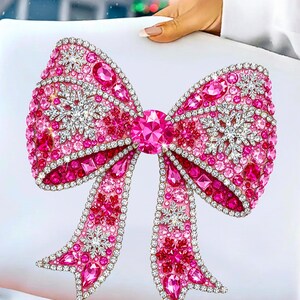 Puede incluir: Un gran lazo rosa decorativo con una joya central y detalles de copo de nieve. El lazo est&aacute; adornado con numerosos cristales rosas y transparentes, creando un efecto brillante. El lazo se sostiene sobre un fondo blanco.