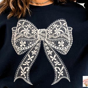Puede incluir: Una sudadera negra con un diseño de lazo de encaje blanco. El lazo es grande e intrincado, con un delicado patrón floral.