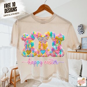 May include: A beige t-shirt featuring three colorful bunny designs with heart-shaped accents and bows. The bunnies are in shades of pink, yellow, blue, and green. The words "happy easter" are embroidered below the bunnies. The text "FREE 10 DESIGNS INCLUDED" is in the upper left corner.