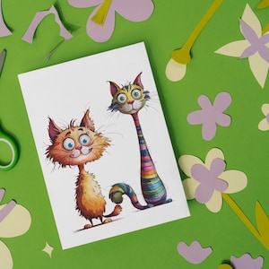 Puede incluir: Ilustraci&oacute;n caprichosa de dos gatos de dibujos animados con ojos grandes y rasgos expresivos. Un gato es naranja, el otro tiene un cuello a rayas arco&iacute;ris. La imagen est&aacute; sobre una tarjeta blanca, rodeada de flores de papel recortadas y tijeras sobre un fondo verde.