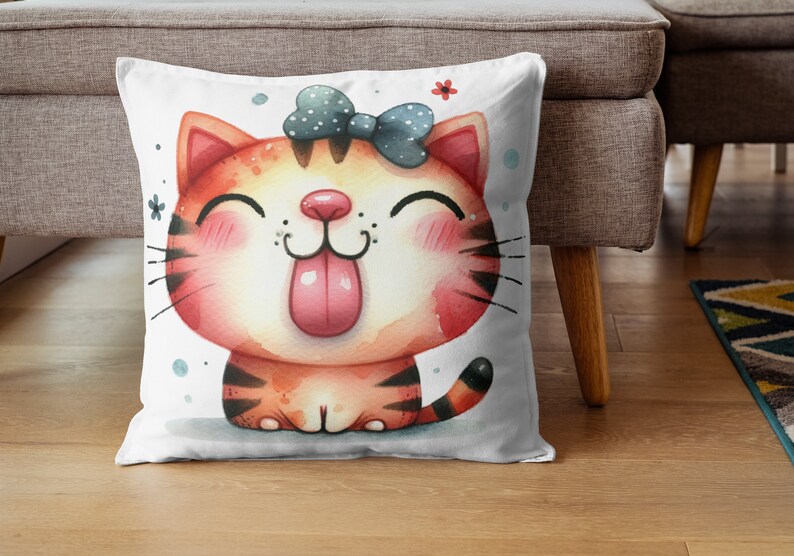 Pode incluir: Uma almofada branca com uma ilustra&ccedil;&atilde;o de desenho animado fofa de um gato tigrado vermelho e laranja com um la&ccedil;o azul. O gato est&aacute; mostrando a l&iacute;ngua e tem uma express&atilde;o feliz.