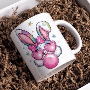 May include: A white ceramic mug with a colorful cartoon bunny wearing pink heart-shaped sunglasses and blowing a pink bubble. The bunny has a pink bow on its head and is surrounded by stars.