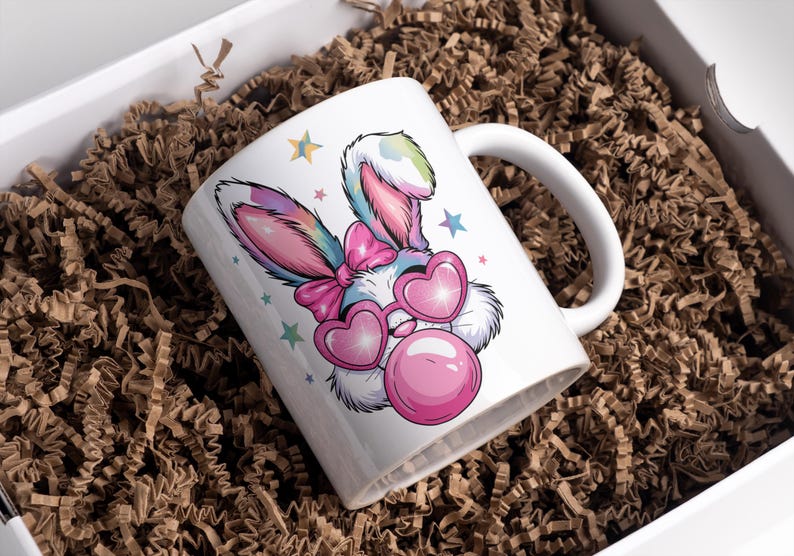 May include: A white ceramic mug with a colorful cartoon bunny wearing pink heart-shaped sunglasses and blowing a pink bubble. The bunny has a pink bow on its head and is surrounded by stars.