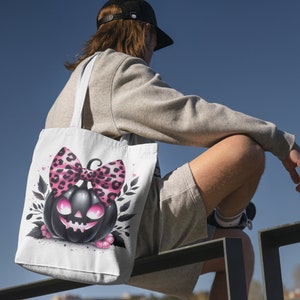 May include: A white tote bag with a black and pink pumpkin with a pink and black leopard print bow. The pumpkin has a spooky face.