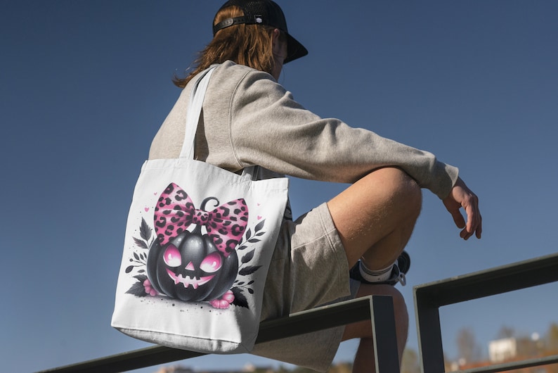May include: A white tote bag with a black and pink pumpkin with a pink and black leopard print bow. The pumpkin has a spooky face.