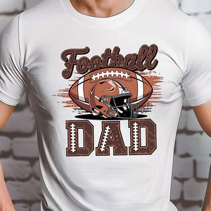 May include: White t-shirt featuring a football design. The design includes a brown football, helmet, and the words "Football DAD" in a brown, distressed font. The shirt is a classic crew neck.