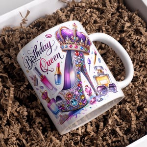 May include: White ceramic mug with a colorful design featuring a high-heeled shoe adorned with jewels, a crown, and makeup items. The text "Birthday Queen" is written in a decorative font.