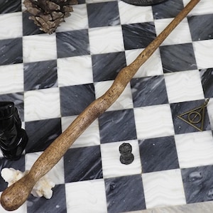 May include: A wooden wand with a curved handle rests on a checkered surface of white and gray squares. A black carved figurine, a pine cone, and a bronze pendant are also visible. The wand is a light brown color.