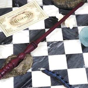 May include: A handcrafted, dark purple wooden wand with a detailed handle rests on a checkered surface. A ticket reading "London to Hogwarts, Platform 9 3/4" lies nearby. Additional elements include a blue-green glass piece and a feather.