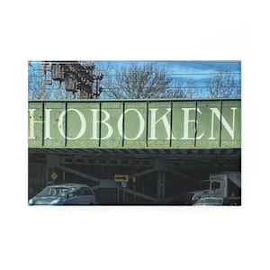 Puede incluir: Un puente de metal verde con la palabra "HOBOKEN" pintada en letras blancas. El puente está sobre una carretera con coches que pasan por debajo.