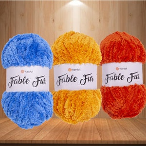 May include: Three skeins of yarn in blue, orange, and yellow. The yarn is fluffy and has a fuzzy texture. The yarn is labeled "Fable Fur" by YarnArt.