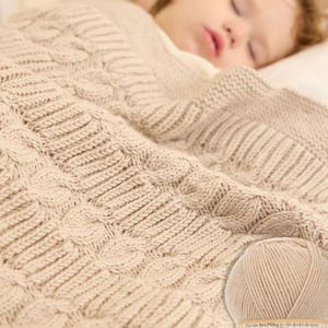 Puede incluir: Una manta de beb&eacute; de punto beige con un dise&ntilde;o de punto trenzado. La manta cubre a un ni&ntilde;o dormido. Se ve un ovillo de lana Alize Baby Best.