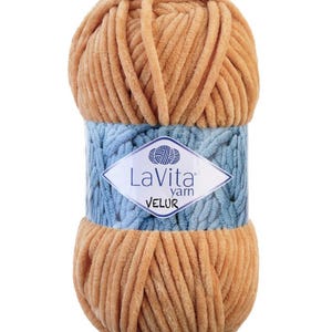 May include: A ball of tan-colored LaVita Velur yarn. The yarn is wrapped with a light blue band featuring the brand's logo. The yarn appears soft and plush, ideal for knitting or crocheting projects. The label indicates it is "Velur" yarn.
