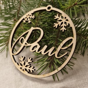 May include: A wooden Christmas ornament with the name "Paul" cut out in a cursive font. The ornament is decorated with snowflakes.