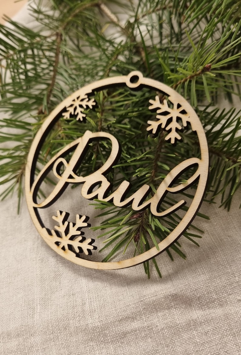 May include: A wooden Christmas ornament with the name "Paul" cut out in a cursive font. The ornament is decorated with snowflakes.