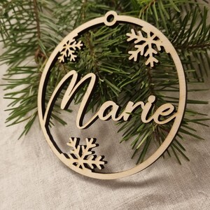 May include: A wooden Christmas ornament with the name "Marie" cut out in the center. The ornament is decorated with snowflakes around the edge.