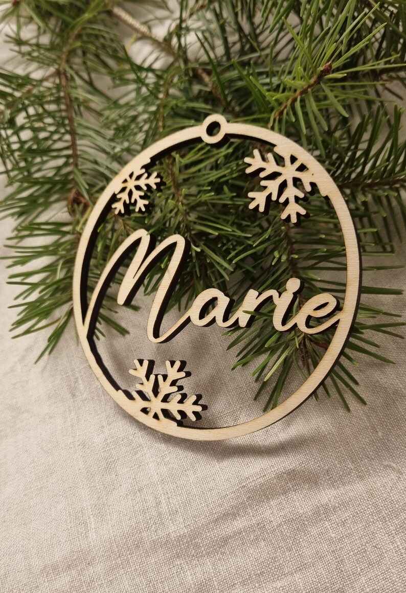May include: A wooden Christmas ornament with the name "Marie" cut out in the center. The ornament is decorated with snowflakes around the edge.