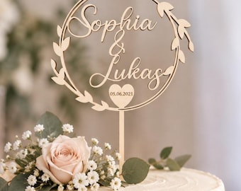 May include: A light-colored wooden cake topper featuring a circular frame adorned with leaf motifs. The center displays the names Sophia and Lukas in cursive script, with a small heart below containing the date 05.06.2021 on a vertical stake.