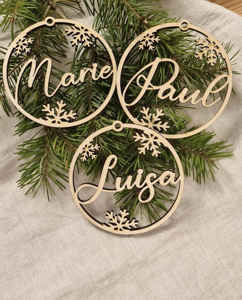 May include: Three wooden Christmas ornaments with the names Marie, Paul, and Luisa written in cursive. Each ornament has a snowflake design and a hole for hanging.