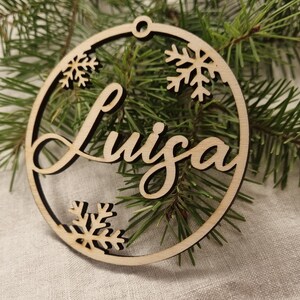 May include: A wooden Christmas ornament with the name "Luiza" written in cursive script. The ornament is decorated with snowflakes.