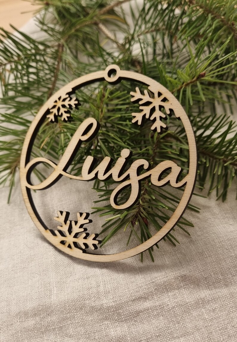 May include: A wooden Christmas ornament with the name "Luiza" written in cursive script. The ornament is decorated with snowflakes.