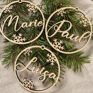 May include: Three wooden Christmas ornaments with the names "Marie", "Paul", and "Luisa" cut out in the center. The ornaments are decorated with snowflakes and are hanging on a green pine branch.