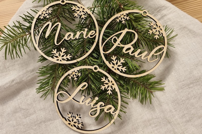 May include: Three wooden Christmas ornaments with the names "Marie", "Paul", and "Luisa" cut out in the center. The ornaments are decorated with snowflakes and are hanging on a green pine branch.