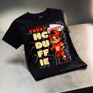 Può includere: T-shirt nero con un'illustrazione a cartoni animati di un giocatore di football americano in uniforme rossa e berretto rosso. La maglietta ha la scritta "TRENT MCDUFFIE CHIEFS" in rosso e giallo. Il giocatore tiene in mano un pallone.