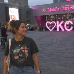Puede incluir: Una camiseta de manga corta de color gris oscuro con un gr&aacute;fico de un bal&oacute;n de f&uacute;tbol dividido en secciones con las banderas de Argentina, Inglaterra, Pa&iacute;ses Bajos y Argelia, junto con el texto KC y una silueta urbana, combinada con vaqueros azules.