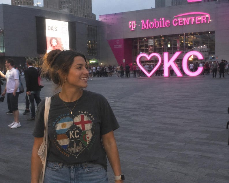 Puede incluir: Una camiseta de manga corta de color gris oscuro con un gr&aacute;fico de un bal&oacute;n de f&uacute;tbol dividido en secciones con las banderas de Argentina, Inglaterra, Pa&iacute;ses Bajos y Argelia, junto con el texto KC y una silueta urbana, combinada con vaqueros azules.