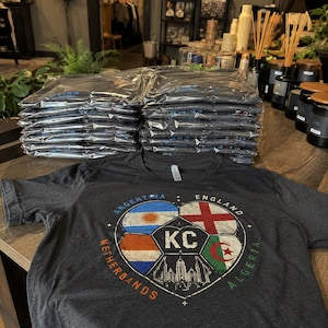 Puede incluir: Una camiseta de manga corta en color gris jaspeado oscuro con un gr&aacute;fico de un bal&oacute;n de f&uacute;tbol dividido en secciones. Las secciones muestran las banderas de Argentina, Inglaterra, Pa&iacute;ses Bajos y Argelia, con las letras KC y un horizonte urbano.
