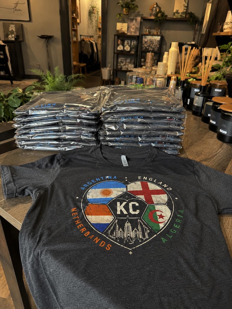 Puede incluir: Una camiseta de manga corta en color gris jaspeado oscuro con un gr&aacute;fico de un bal&oacute;n de f&uacute;tbol dividido en secciones. Las secciones muestran las banderas de Argentina, Inglaterra, Pa&iacute;ses Bajos y Argelia, con las letras KC y un horizonte urbano.