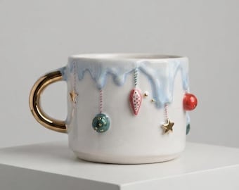 May include: A white ceramic mug with a shiny gold handle, featuring a light blue drip pattern around the rim. The mug is decorated with small, raised, colorful ornaments including red and green spheres, a pink teardrop shape, and gold stars.