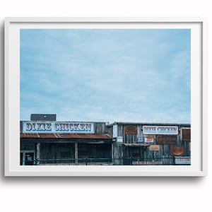 以下が含まれることがあります： 「Dixie Chicken」の文字が入った看板がある、木造の素朴な建物の額装写真。建物は風化した外観で、青い空を背景にしています。