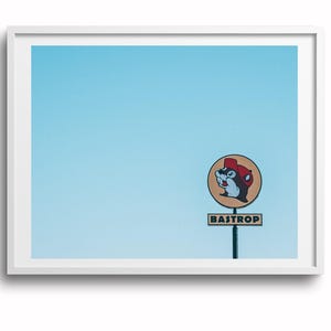 May include: A framed print featuring a sign with a cartoon beaver wearing a red hat, set against a clear, light blue sky. The sign reads "BASTROP" in a rectangular box below the beaver graphic.