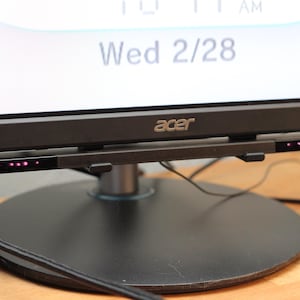Op de afbeelding: Een zwarte Acer-monitor met een zwarte standaard en een zwarte sensorbalk met een rij roze lampjes. De monitor toont de datum "Wed 2/28".