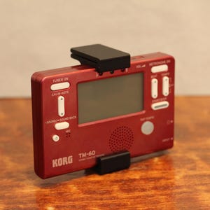 May include: Red Korg TM-60 chromatic tuner and metronome with a black clip. The tuner has a digital display, buttons for tuner on, calib-note, sound in, sound back, and mic. The metronome has buttons for beat, tempo, and tap tempo.